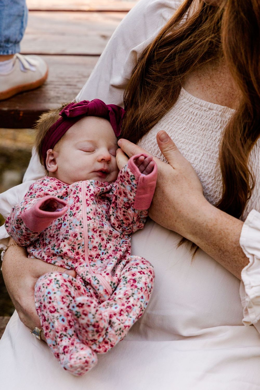 A sleeping baby wearing a pink floral outfit rests peacefully against someone in a white sweater.