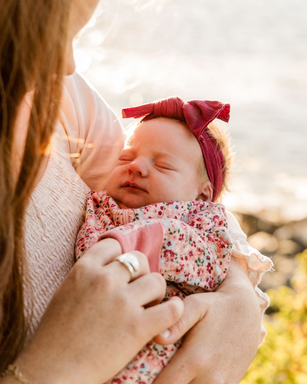 Baby wearing floral outfit and burgundy bow rests peacefully in arms during outdoor sunset photoshoot.