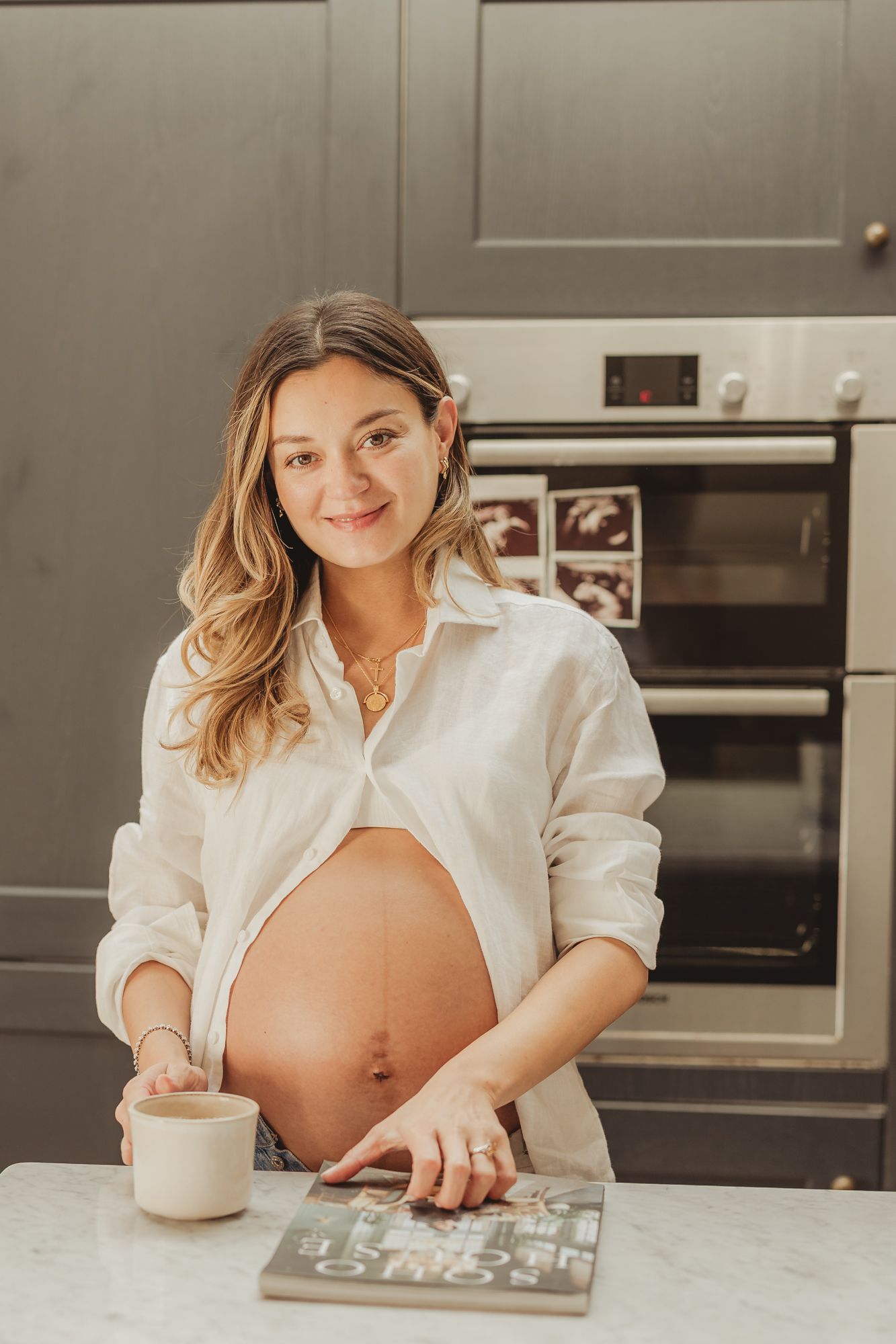A pregnant woman in a white button-down shirt sits at a kitchen counter looking at a photo album.
