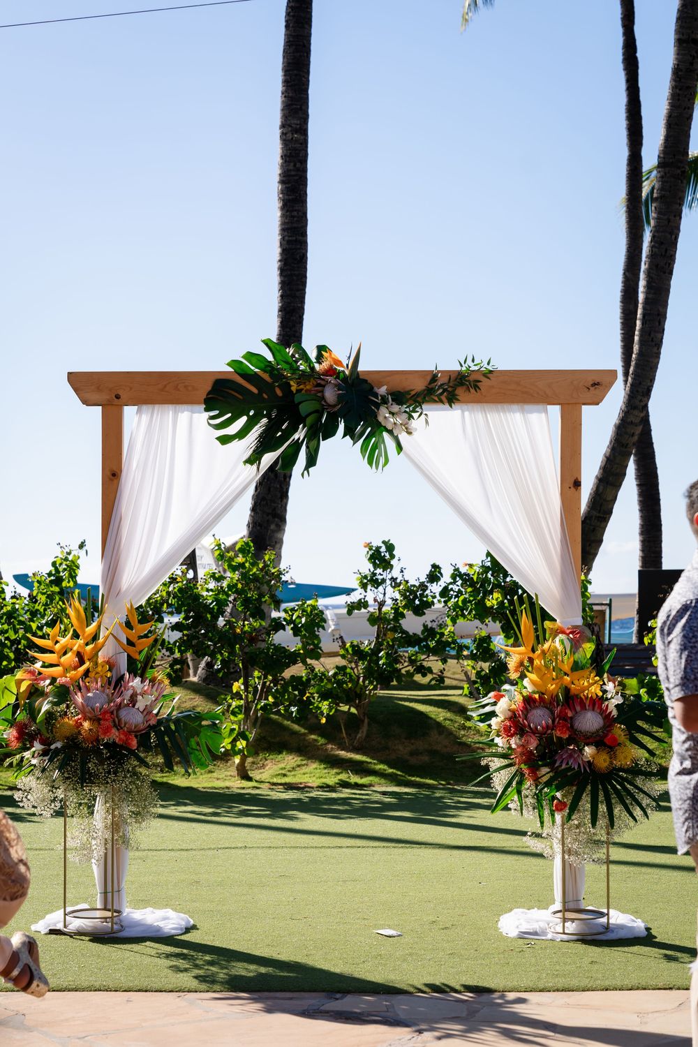 Tropical beach wedding setup with white curtained pergolas and vibrant floral arrangements against palm trees and ocean view.