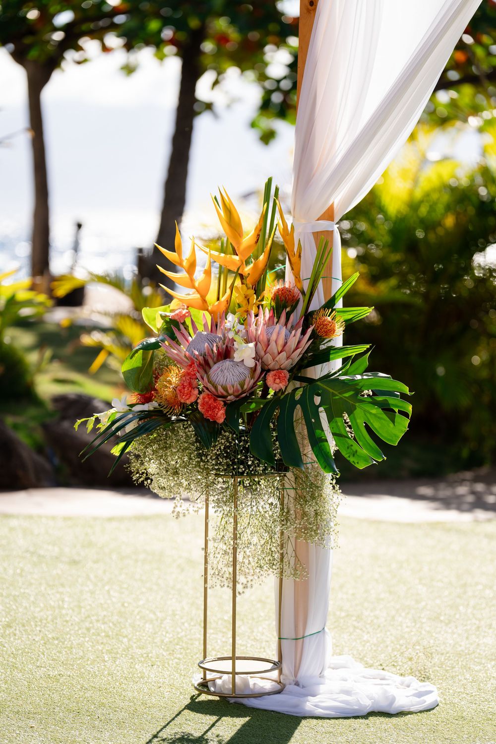 Tropical floral arrangement with proteas and palm leaves adorns white fabric draping at an outdoor wedding ceremony.