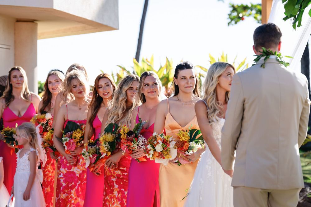 Wedding party with bridesmaids in bright coral dresses and groomsmen in light suits stand together in outdoor venue.