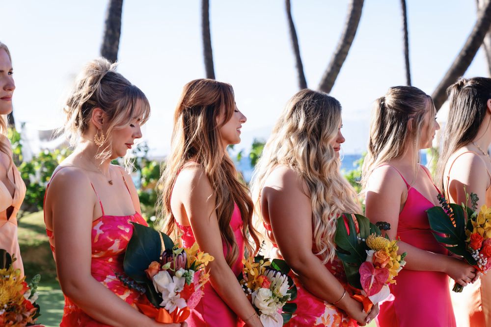 Row of bridesmaids in coral dresses holding colorful bouquets against a tropical backdrop.