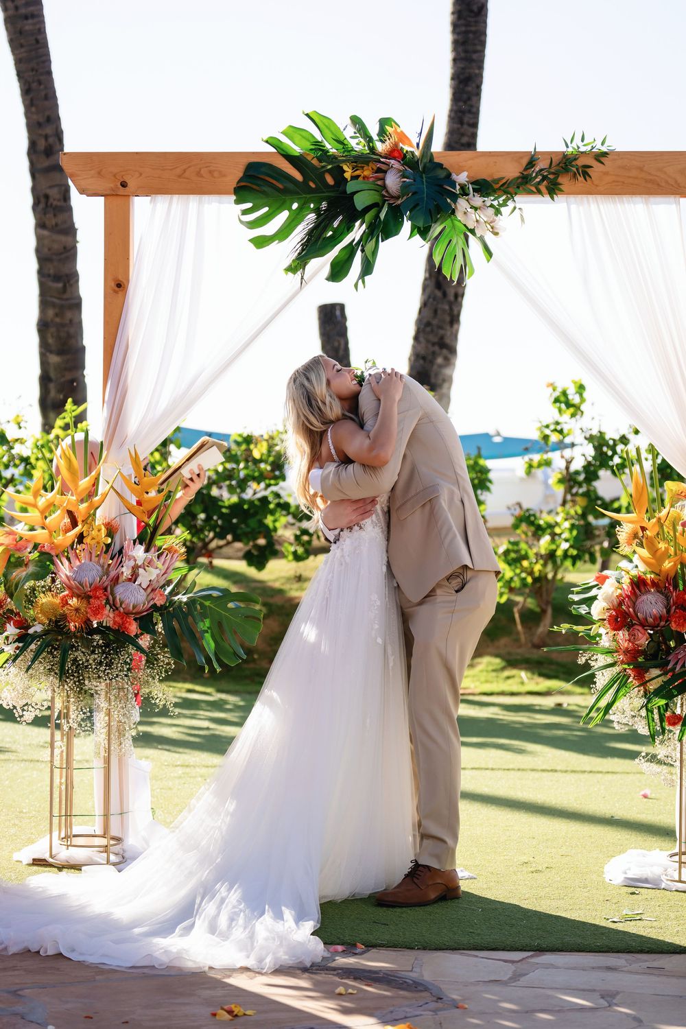 A wedding couple embraces during their tropical beachside ceremony with palm trees and floral decorations.