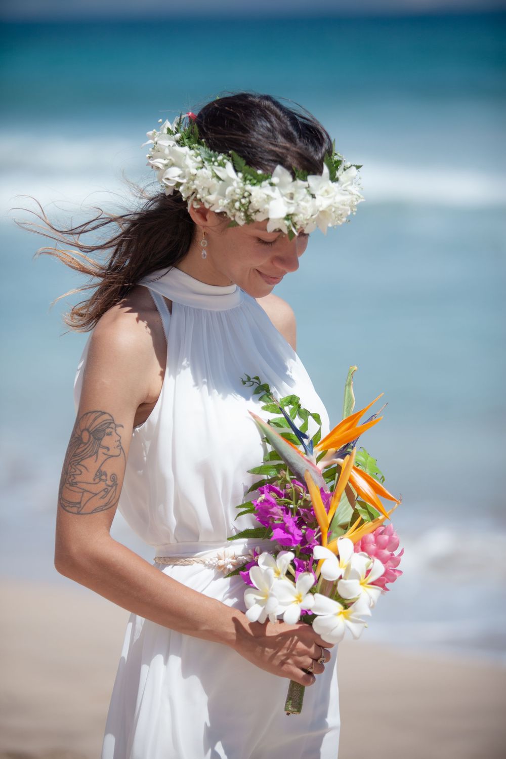 Someone wearing a white dress holds a vibrant tropical bouquet featuring birds of paradise and orchids on a beach.
