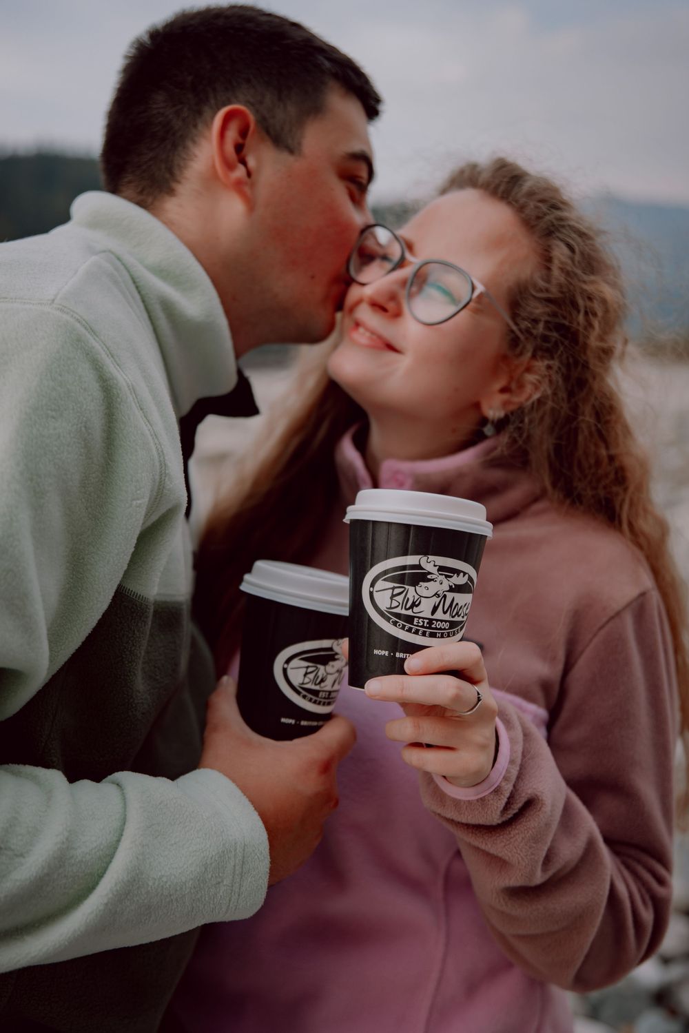 A couple shares intimate moments while holding coffee cups during an outdoor photoshoot.