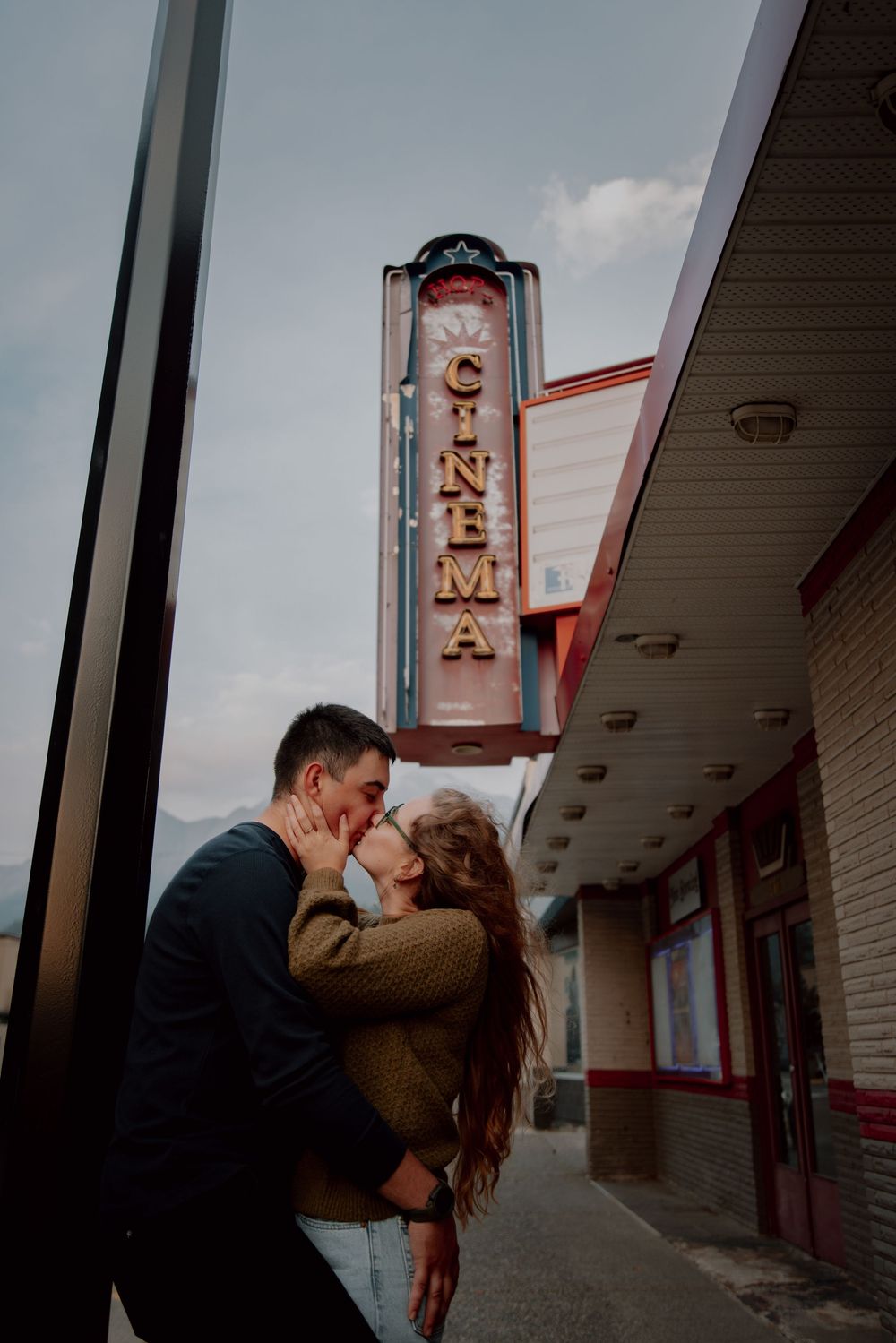 A romantic couple shares a tender kiss outside a vintage movie theater with neon signage at dusk.