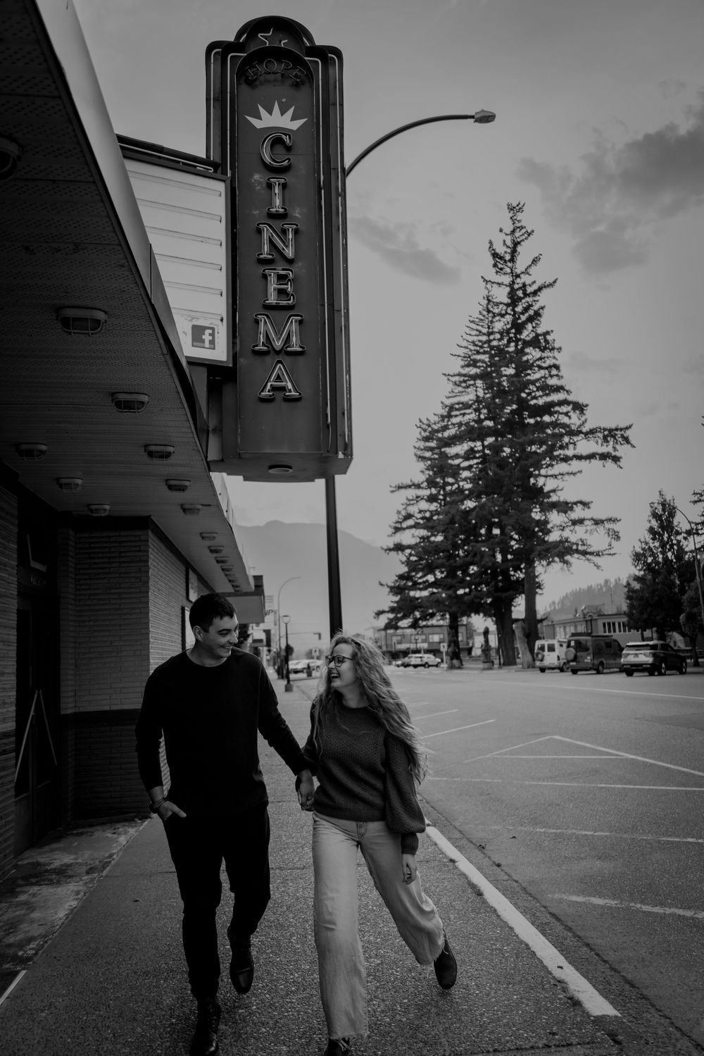 A couple walks playfully in a black and white scene outside a cinema with vintage signage and evergreen trees.