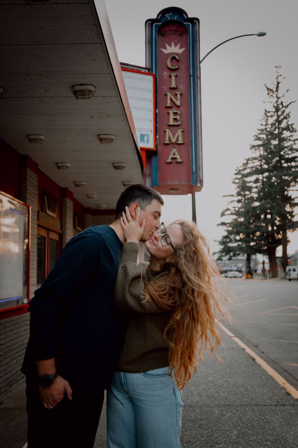 A romantic couple embraces and kisses outside a vintage cinema theater on an overcast evening.