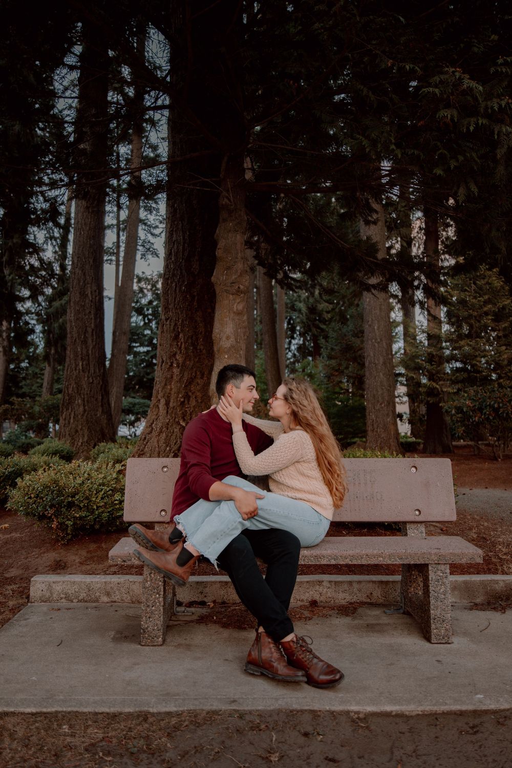 A romantic couple sits and embraces on a stone bench surrounded by tall pine trees in a forest setting.