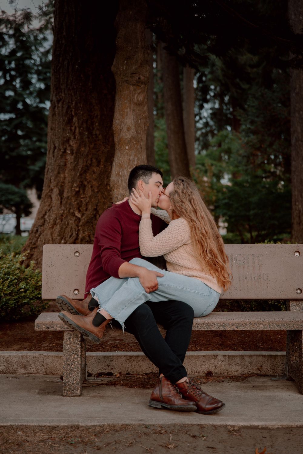 A couple shares intimate moments while sitting together on a concrete bench beneath tall trees in a park setting.