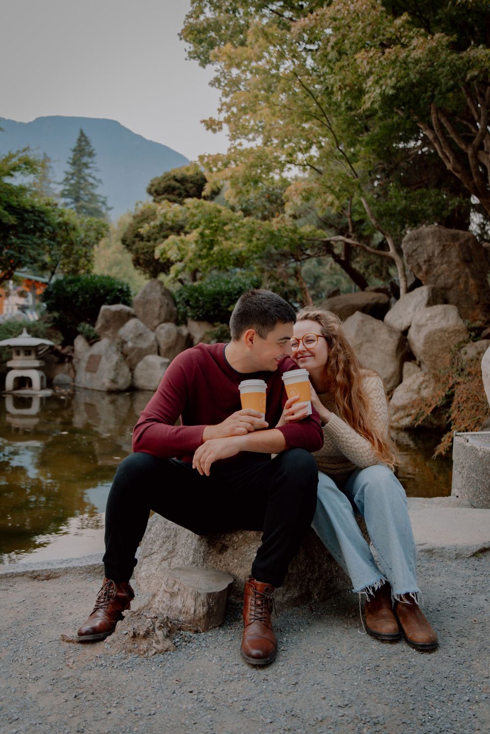 A couple sits together on rocks near a serene pond in a romantic outdoor setting at dusk.
