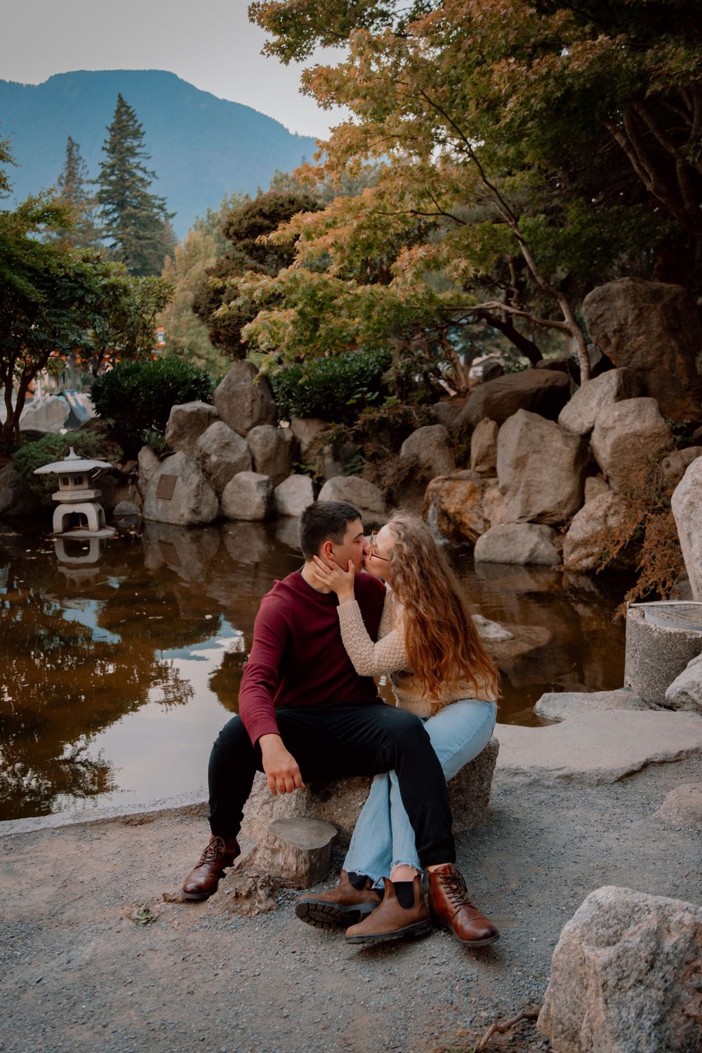 A couple shares intimate moments while sitting on rocks near a tranquil pond in a Japanese garden setting.