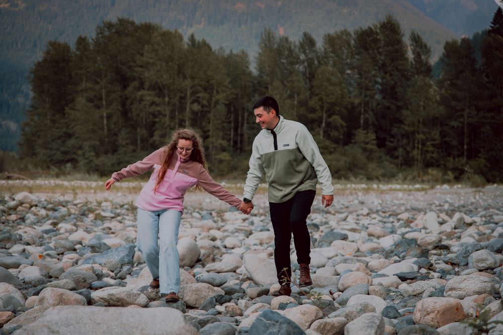 Two people holding hands while carefully walking across a rocky riverbed surrounded by evergreen forest.