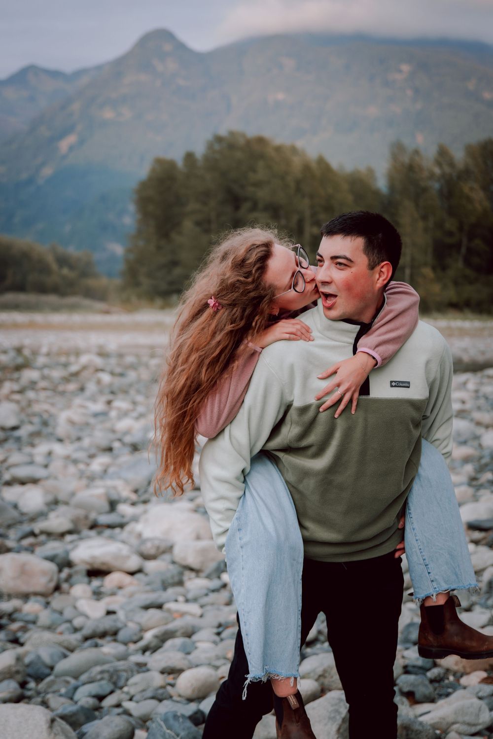 A romantic couple shares playful moments on a rocky riverbed with mountains in the background.