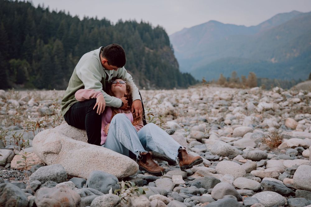 A couple shares an intimate moment on a rocky riverbed with mountains and forest in the background.