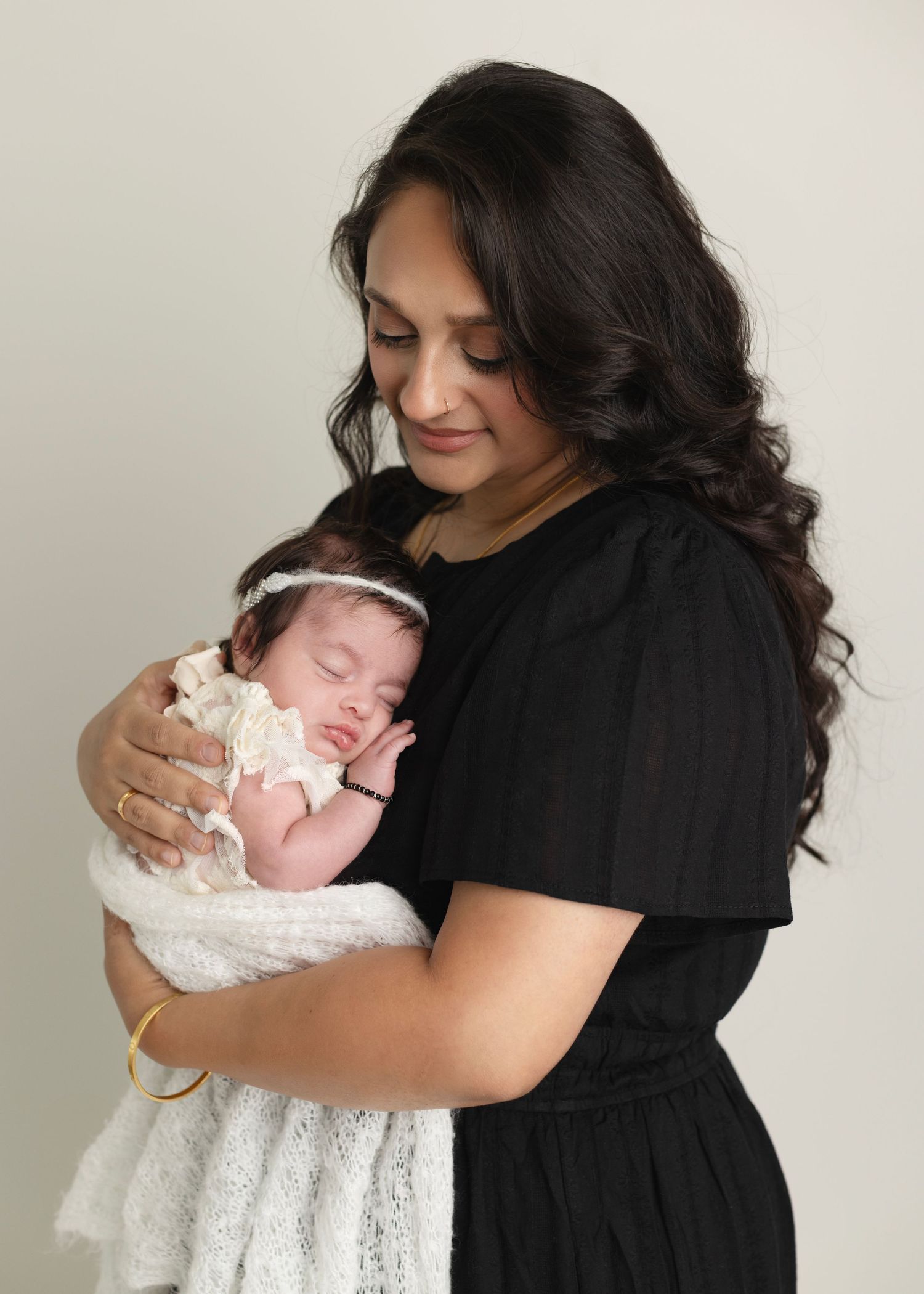 A woman in a black dress holds and gazes lovingly at a sleeping newborn baby wrapped in white.