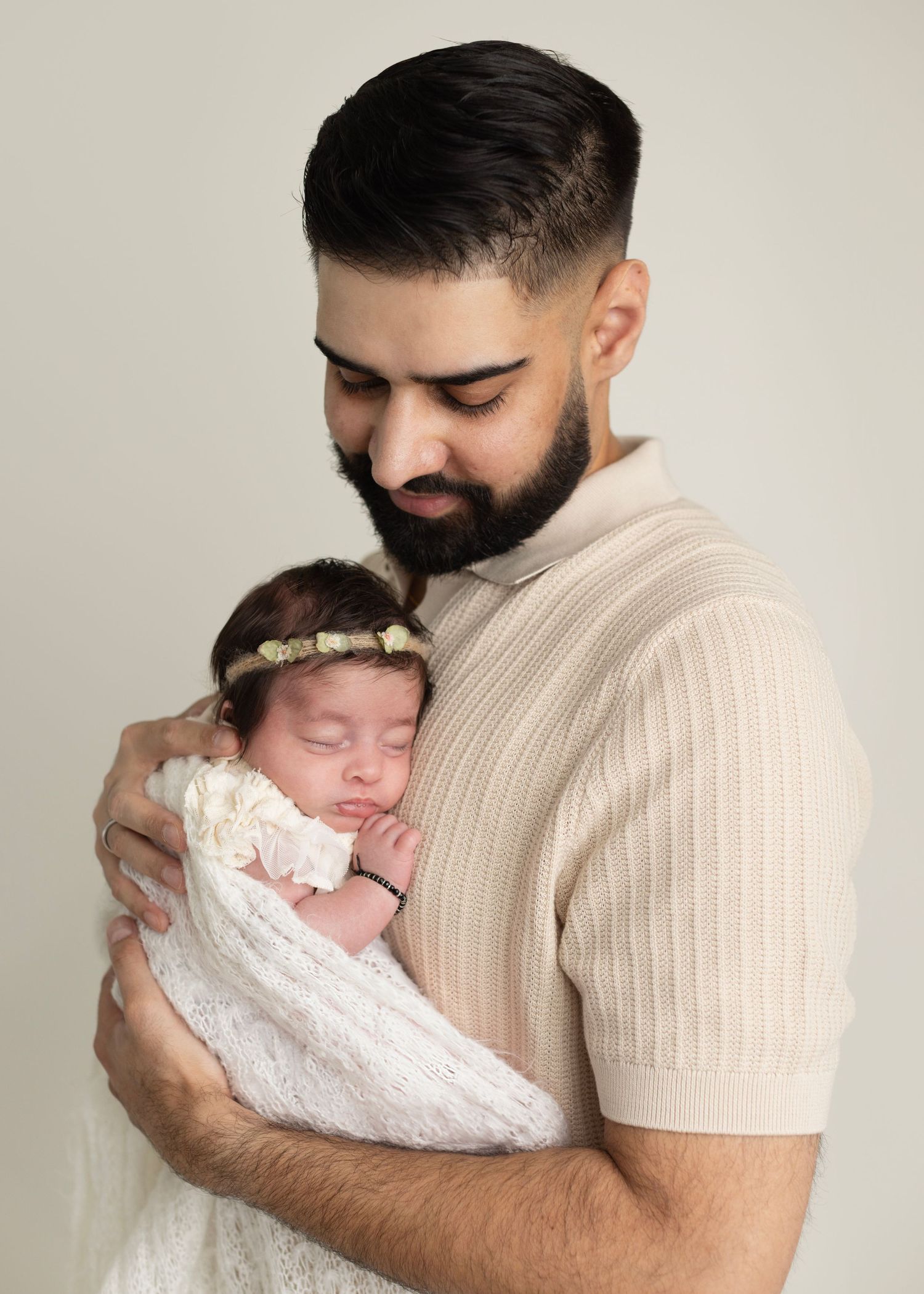 A tender moment captured as a bearded person in a beige sweater cradles a sleeping newborn wrapped in white lace.
