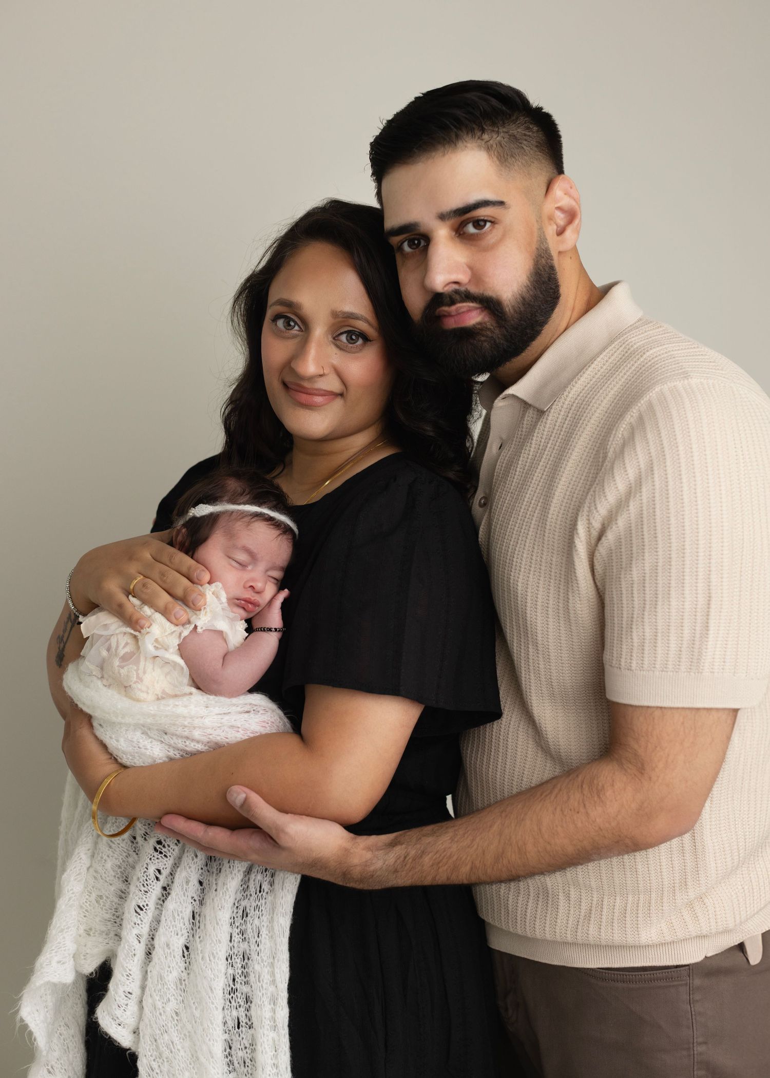 A couple embraces while holding a newborn baby wrapped in white against a neutral grey background.