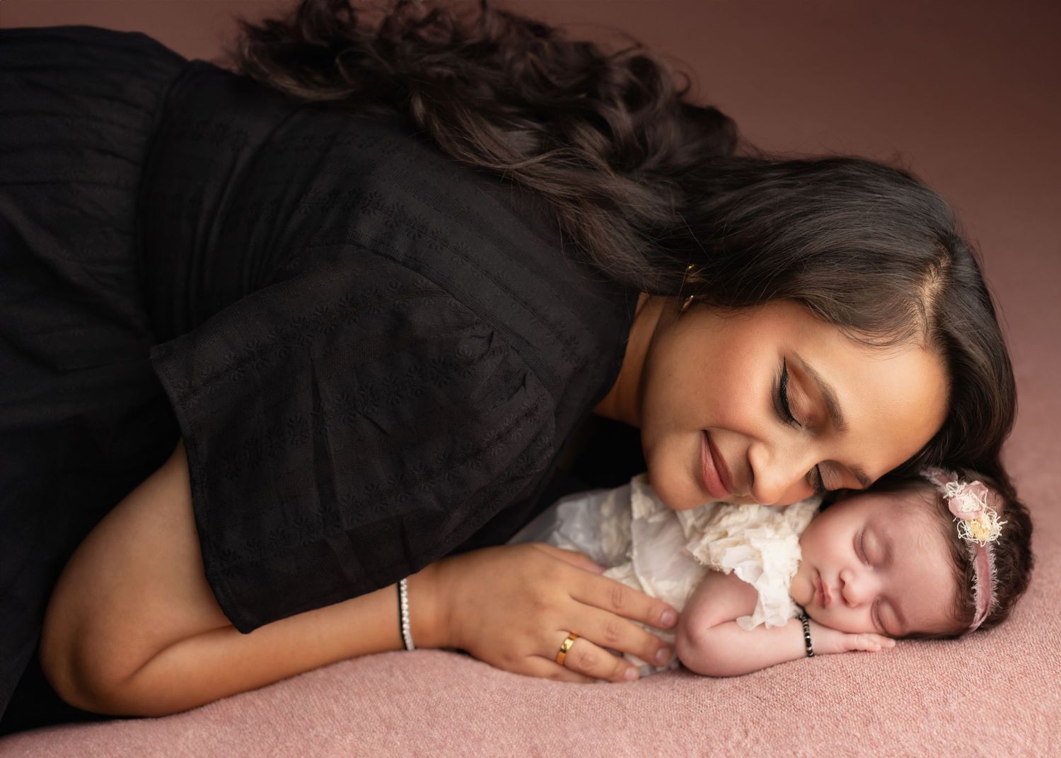 A tender moment captured on pink fabric as a mother in black gently cuddles with her sleeping newborn baby.