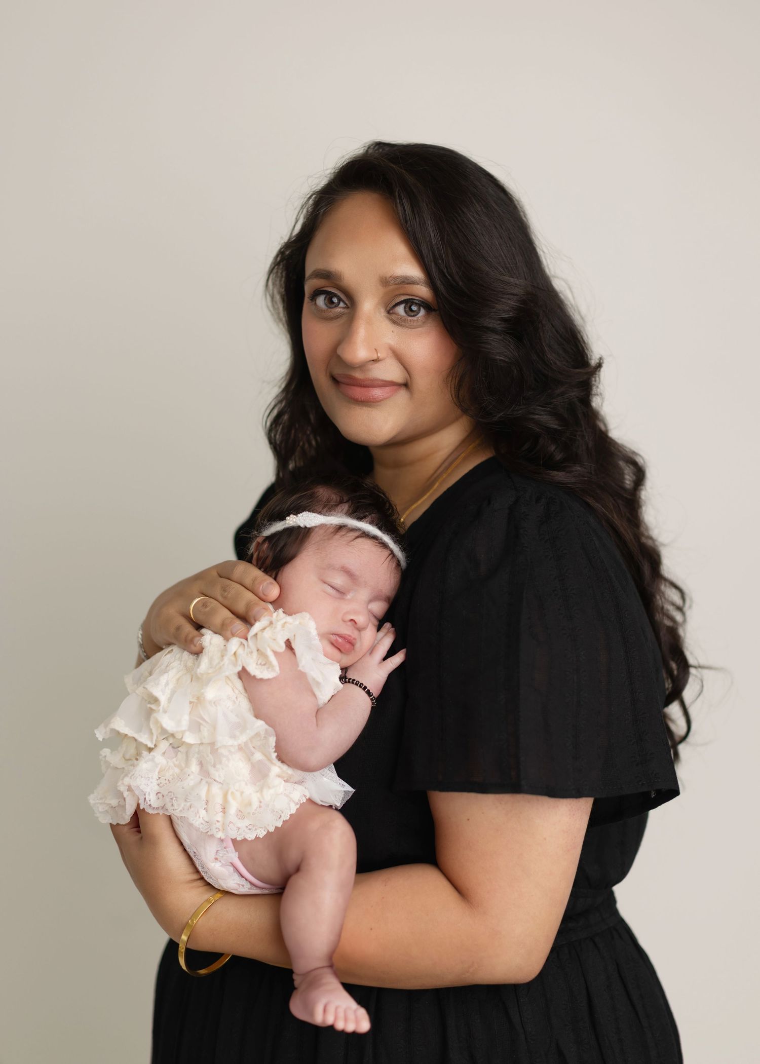 A woman in a black dress holds a sleeping baby wearing a white ruffled outfit against a neutral background.
