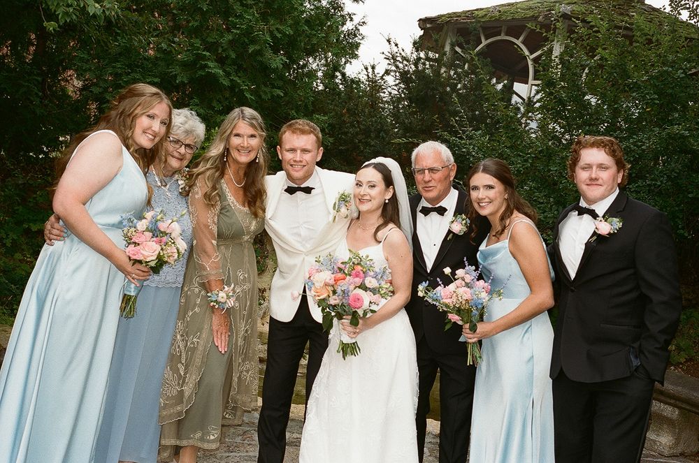 Wedding party in light blue bridesmaid dresses and dark suits pose together in a garden setting.