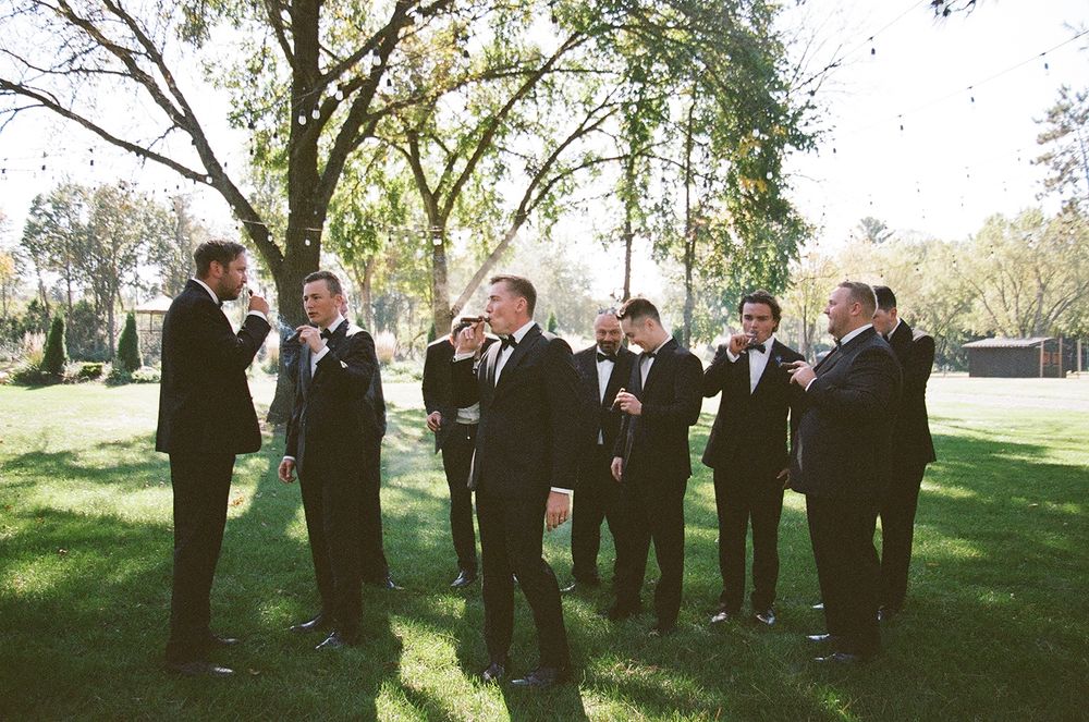 Groomsmen in black tuxedos pose together under trees at an outdoor wedding ceremony.