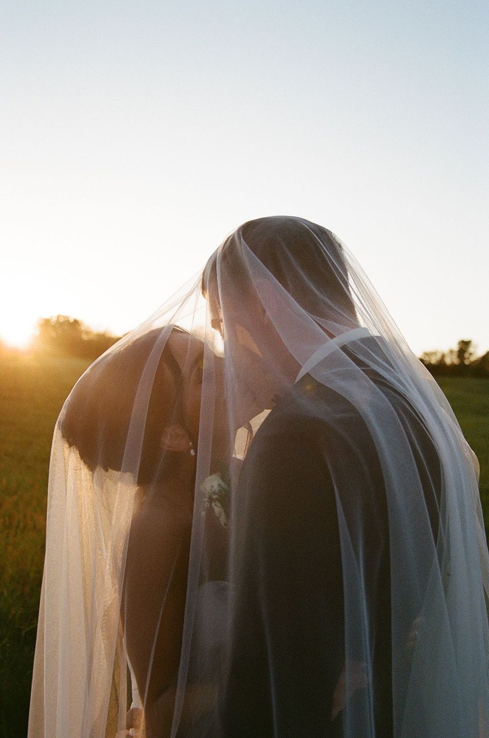 Two people in wedding attire share a romantic moment under a flowing veil at sunset in a field.