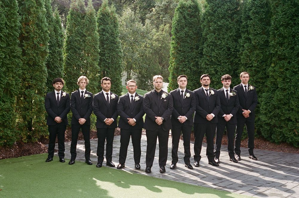 Wedding party in black suits stands in line against green hedge backdrop in outdoor garden setting.