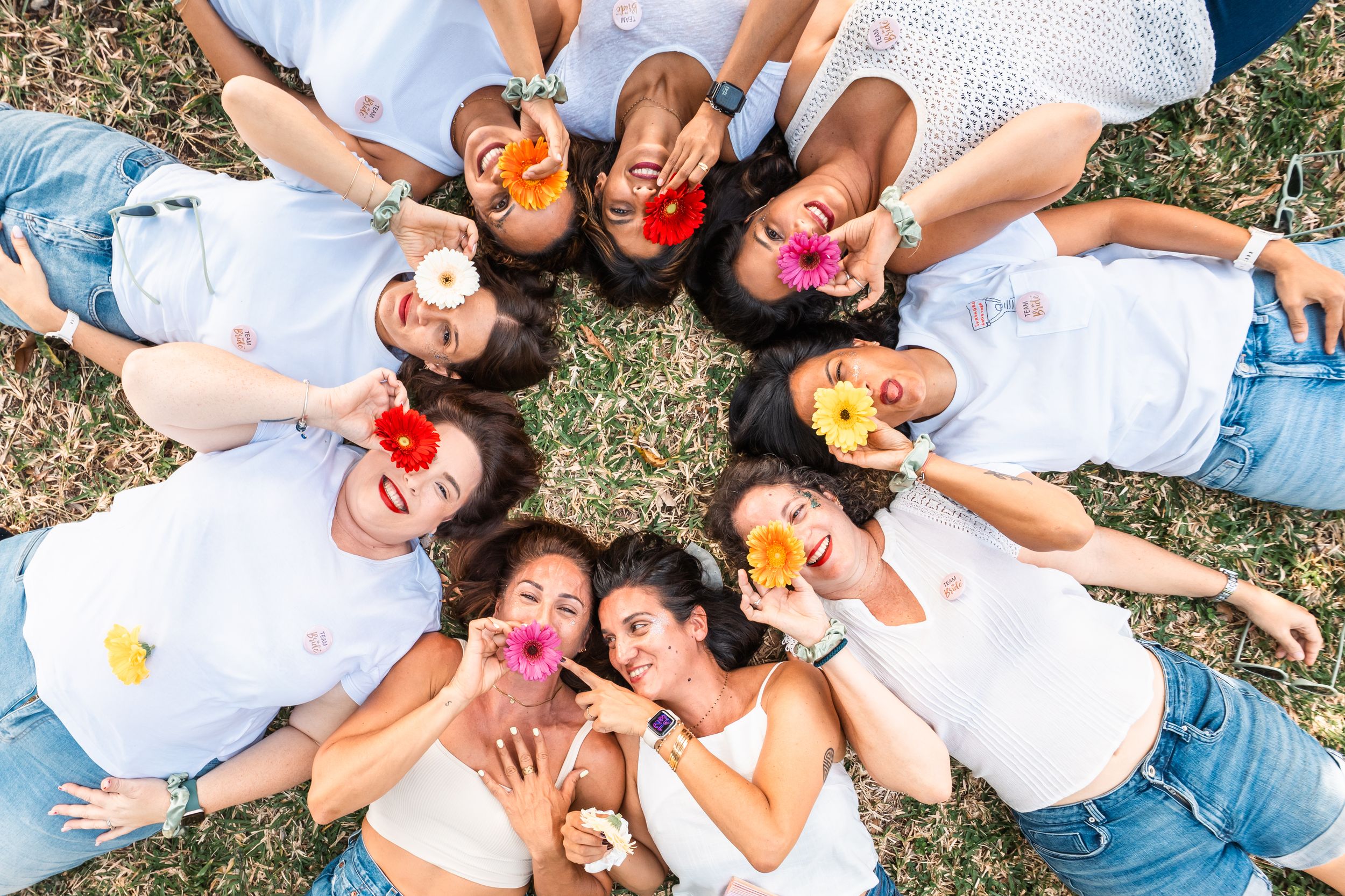 A group of friends lying in grass arranged in a circle wearing white shirts and holding colorful drinks.
