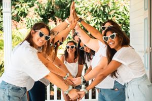 Friends in white tops and denim make heart shapes with their arms while laughing in a sunny outdoor setting.
