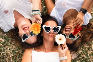 A group lying on grass wearing white outfits and heart-shaped sunglasses while holding colorful gerbera daisies.