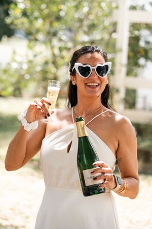 Person in white dress and heart-shaped sunglasses enjoying champagne on a sunny day outdoors.