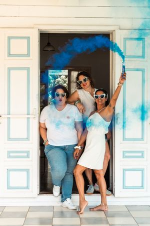 Group poses playfully with colorful blue smoke against white building exterior.