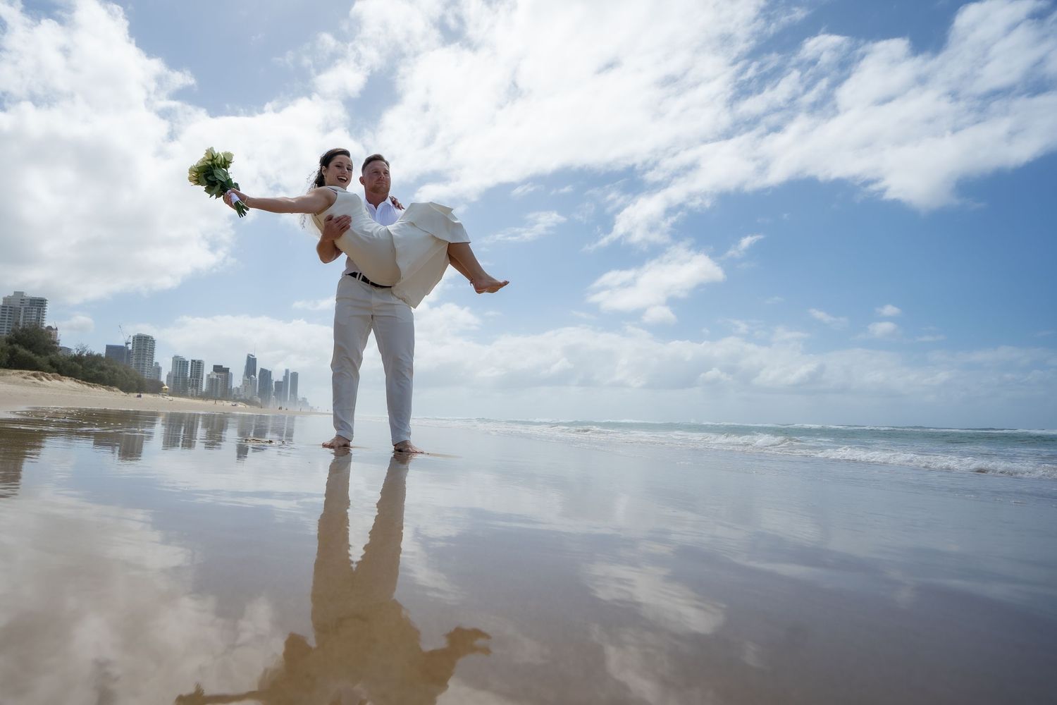A couple in white outfits playfully poses on a sunny beach with ocean waves and city skyline in the background.