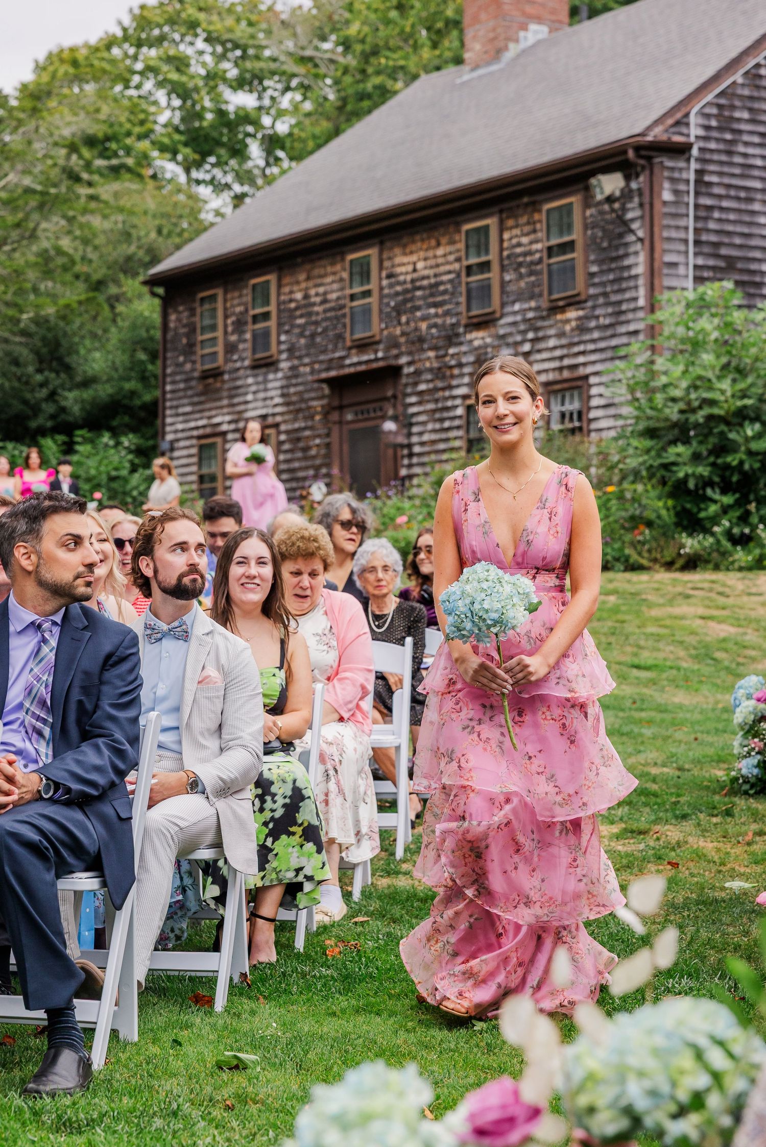 Wedding guests seated on white chairs watch as someone in a pink ruffled dress walks down the aisle at a rustic outdoor ceremony.