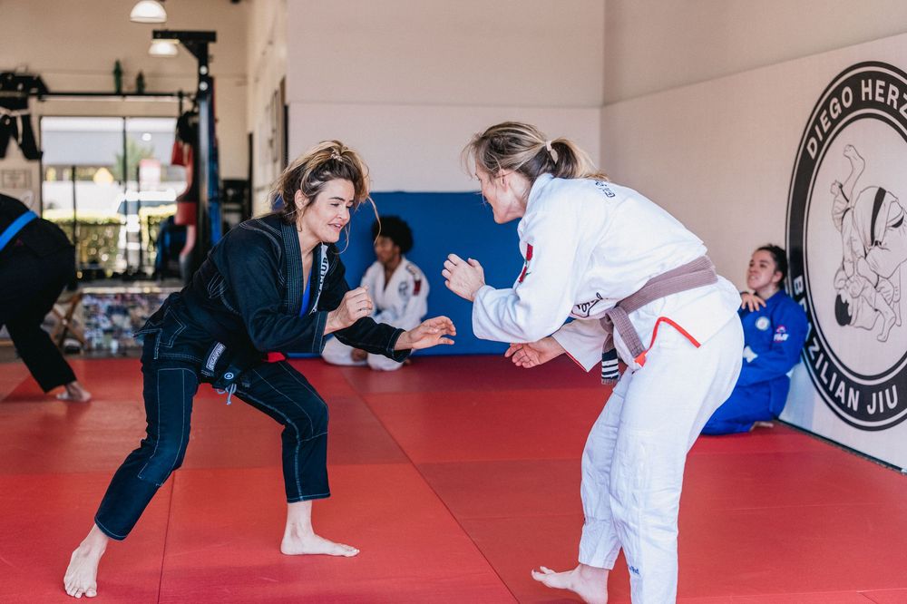 Two martial artists practicing jiu-jitsu techniques on red mats in a training facility.