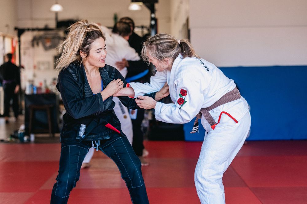 Two people demonstrating martial arts moves and grappling techniques on red mats in a training facility.
