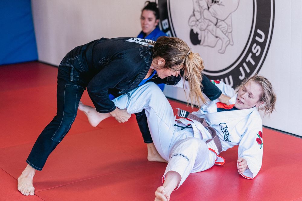Two martial artists practicing grappling techniques on a red mat during a Brazilian Jiu-Jitsu training session.