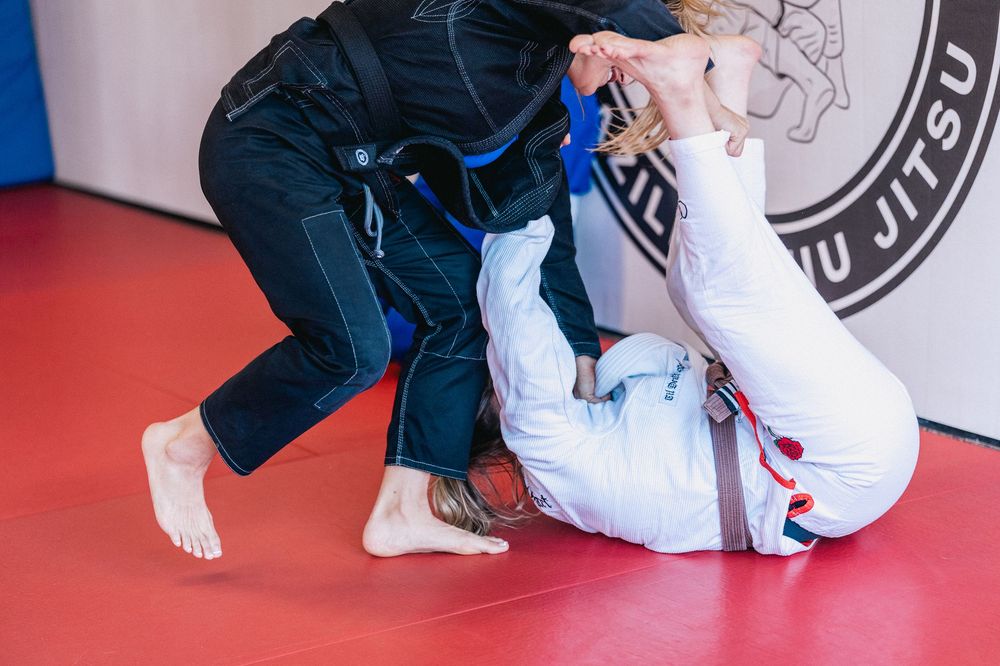 Two martial artists practicing Brazilian Jiu-Jitsu grappling techniques on red and blue mats during training.