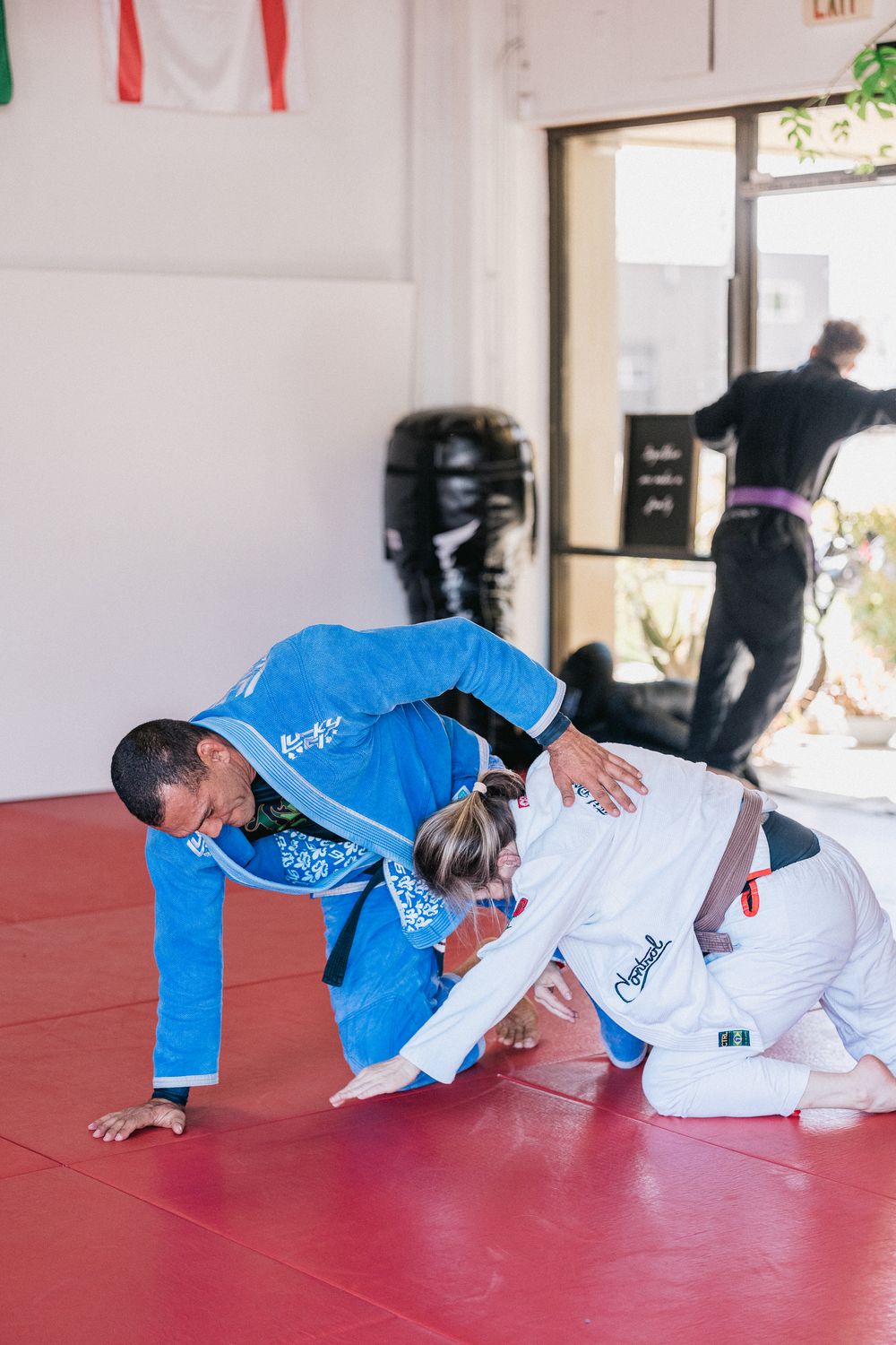 Two martial artists in white and blue gis demonstrate a grappling technique sequence on red training mats.