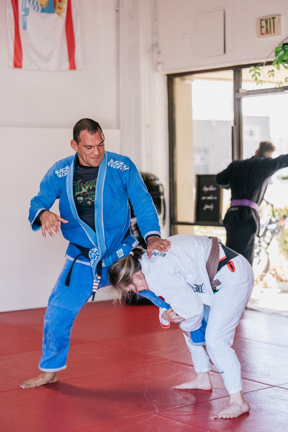 A martial arts instructor demonstrates a judo or jiu-jitsu takedown technique sequence on a red training mat.
