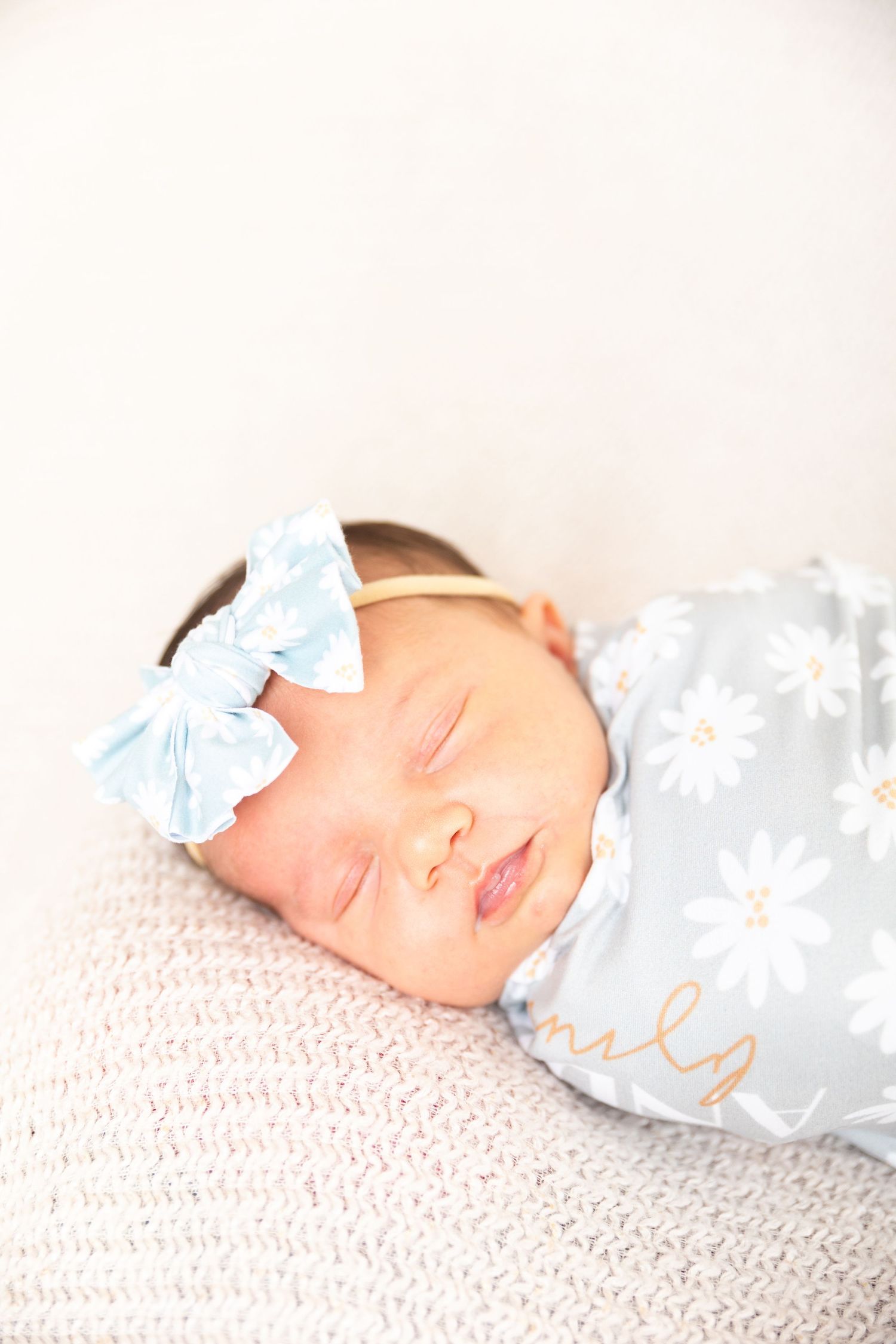 A sleeping newborn baby wearing a light blue bow rests peacefully on textured white bedding.
