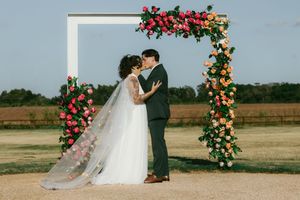 A floral arch decorated with vibrant pink and orange blooms frames an outdoor wedding ceremony in a rustic field setting.
