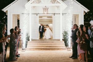 A nighttime wedding ceremony exit at a white chapel with illuminated columns and guests lining the walkway.
