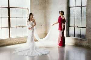 A bride in a white lace wedding gown with a flowing train stands by a window as someone in red helps adjust the dress.