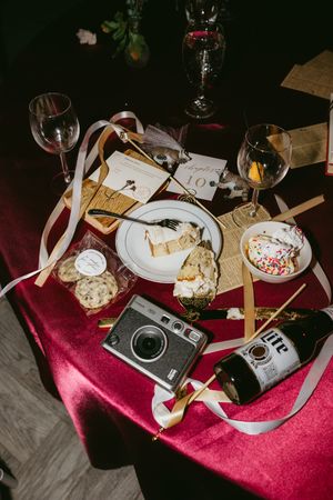 Vintage cameras and wine glasses arranged on red velvet tablecloth with cheese plate and scattered ribbons.