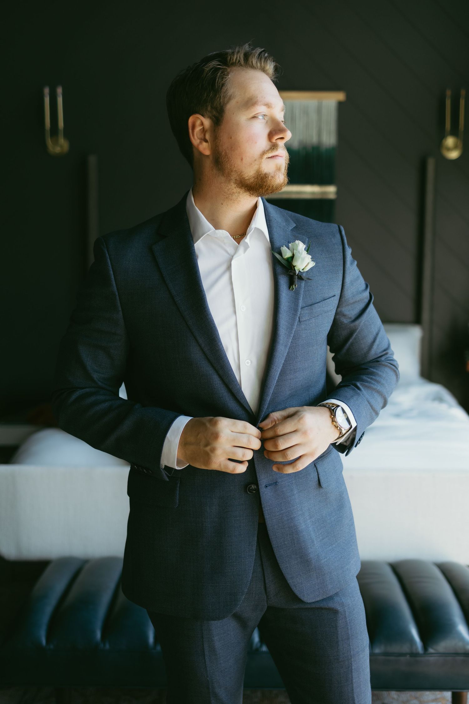 A groom in a navy blue suit adjusts his jacket while standing in a dimly lit room with a boutonniere on his lapel.