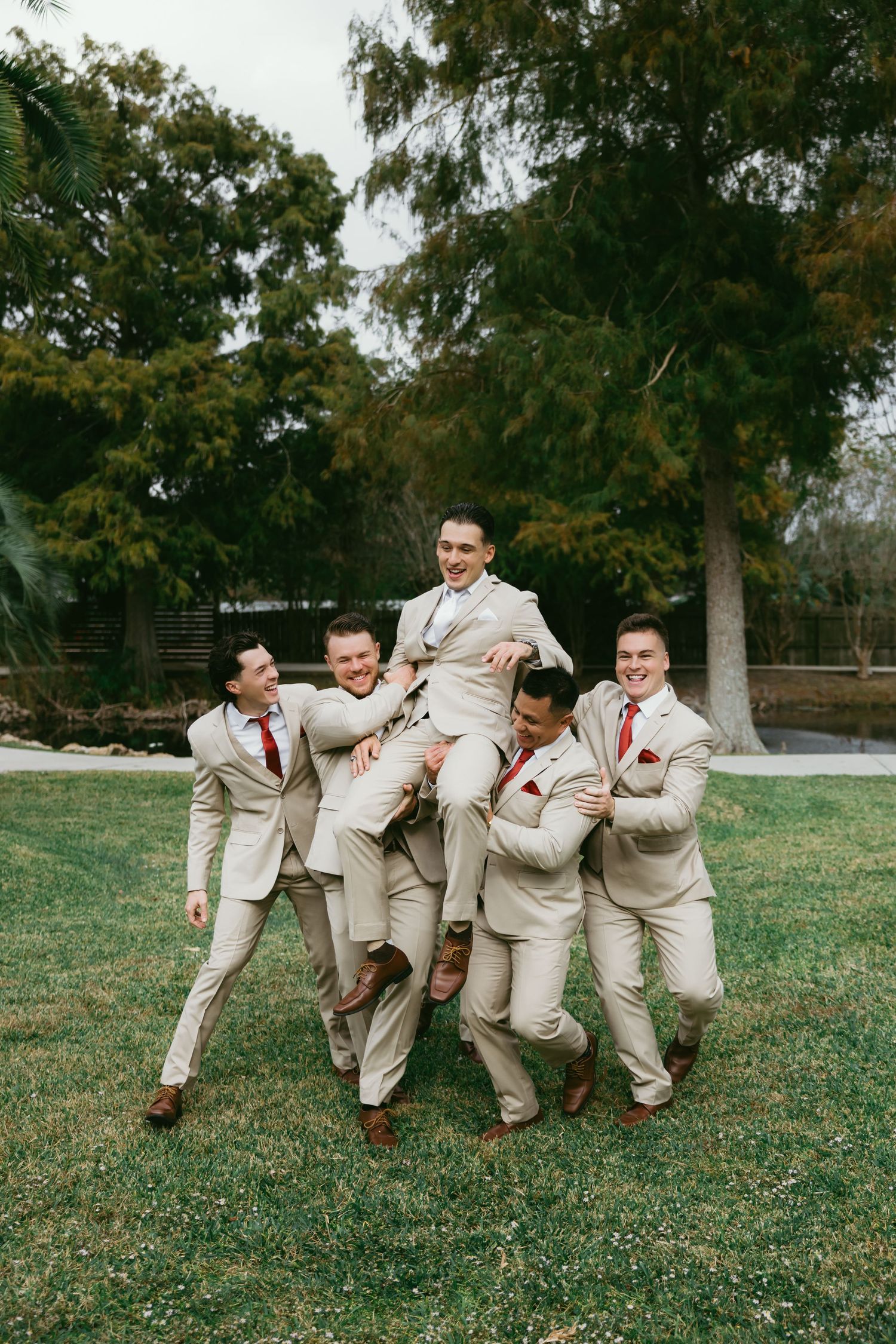 Groomsmen in beige suits perform a domino effect falling pose on green grass during a wedding photoshoot.