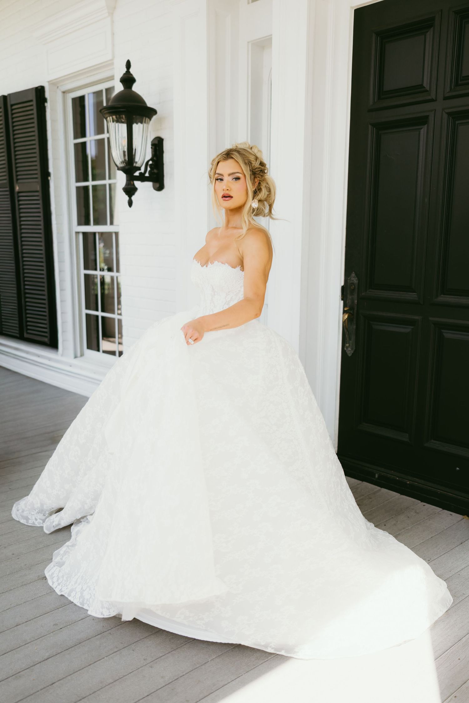 A stunning white wedding dress with a flowing train is showcased on a classic white porch with black shutters.
